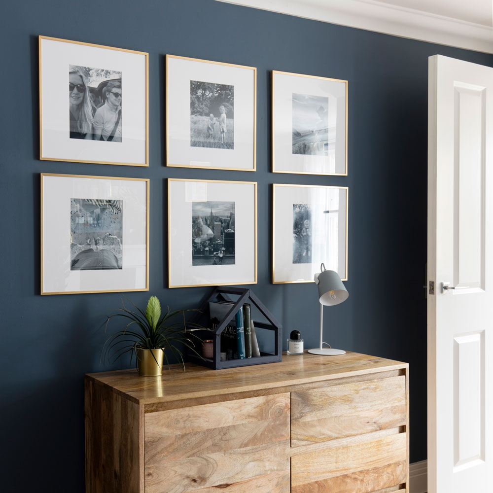 Pictures in gold frames against blue painted wall above wooden chest of drawers in bedroom
